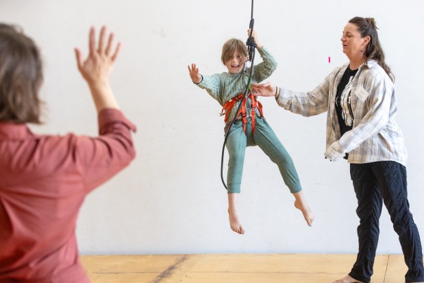 OAKLAND, CA - NOVEMBER 12: Avery Smith waves to her mother Taylor Samuelson while hanging from a rope attached to a harness during a flight test vertical dance lesson under the instruction of Sarah Keeney (right) at the BANDALOOP open house at their studio space in Oakland, Calif. on Nov. 12, 2023. BANDALOOP is a renown vertical dance troupe, that have performed their aerial mid-air performances on international landmarks around the world. (Douglas Zimmerman/Special to the Bay Area News Group)